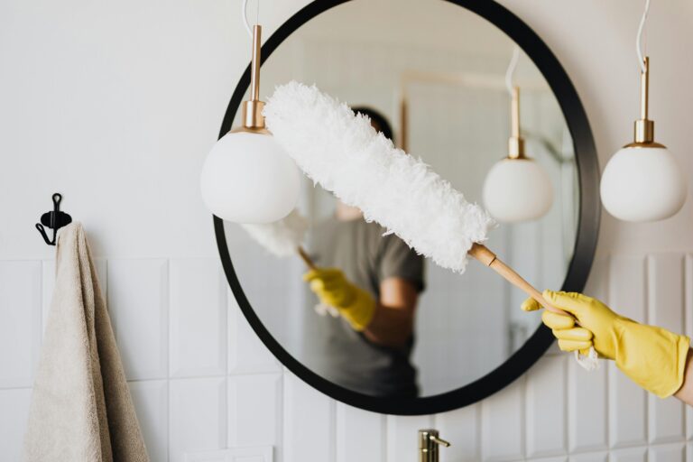 A person cleaning an elegant bathroom mirror with a fluffy duster and yellow gloves.