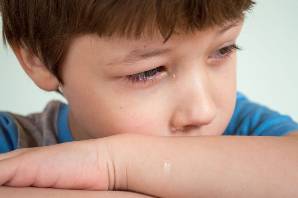 Portrait of crop sad child with brown hair and tears on face crying and lying on arms while looking away against white background