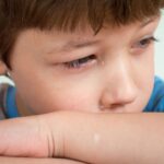 Portrait of crop sad child with brown hair and tears on face crying and lying on arms while looking away against white background